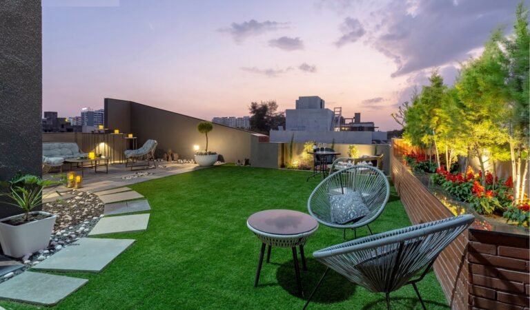 Rooftop garden at dusk with modern furniture, green artificial turf, plants, and city skyline.