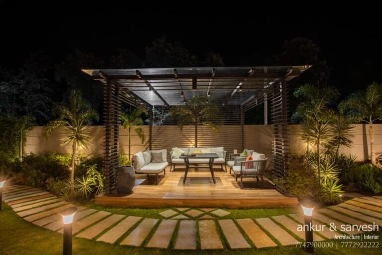 An elegantly lit outdoor seating area with modern furniture under a pergola, surrounded by lush plants at night.