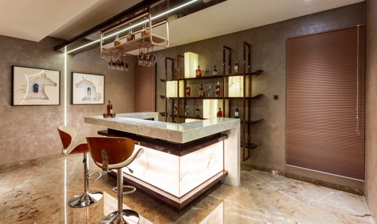 Modern home bar with backlit marble counter, wooden stools, and wall-mounted shelving with bottles.