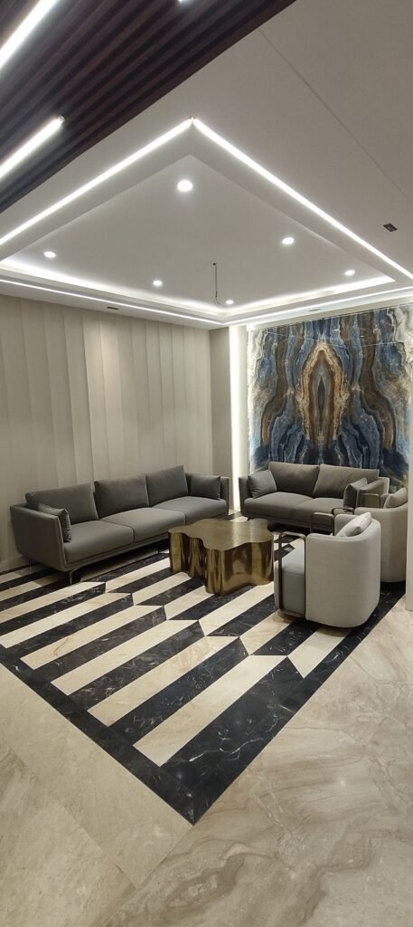 Luxurious office interior design in Gurgaon featuring modern grey seating, abstract onyx wall panel, bold striped marble flooring, gold table, and recessed LED lighting.