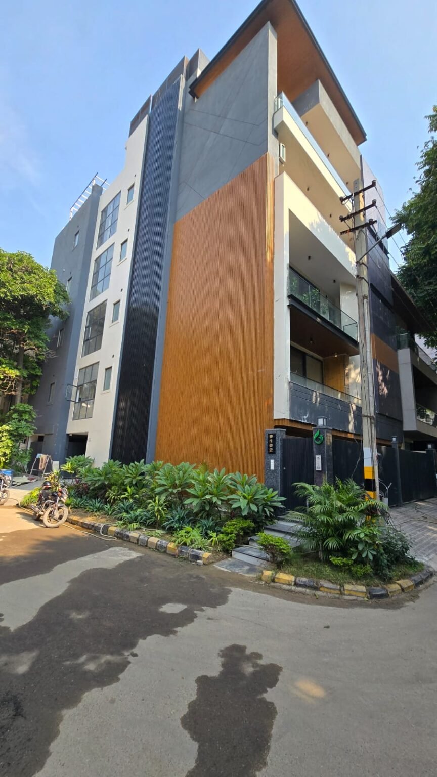 Modern multi-storey building with wooden and black panel accents, showcasing modern interior design services in Gurgaon with glass balconies and lush entrance landscaping.